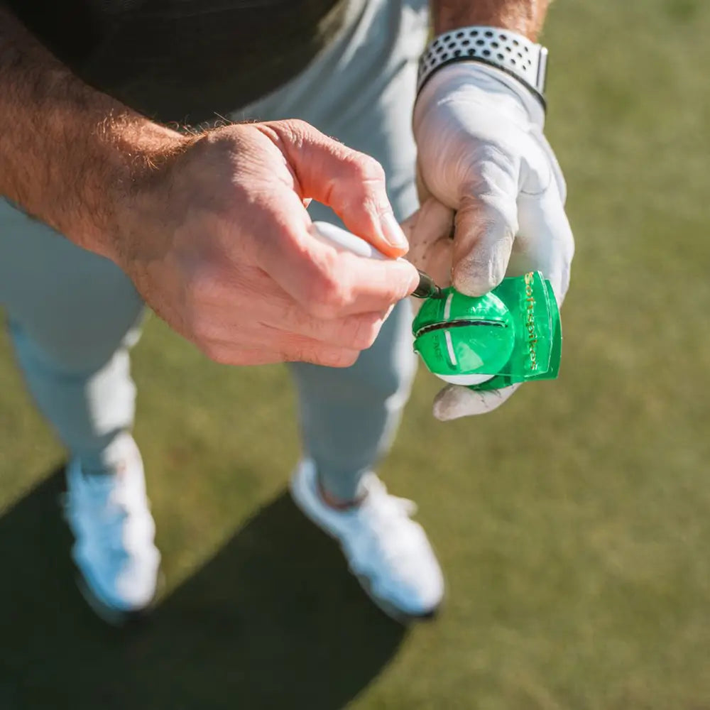 Person using green alignment tool on a golf ball with a white glove on a golf course.
